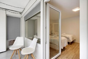 Quarto com porta de vidro deslizante, duas cadeiras brancas, cama com roupa de cama branca e piso de madeira.