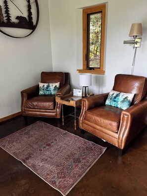 A room with two leather armchairs, a small wooden table, a rug, a wall-mounted lamp, and a window with a view of trees.