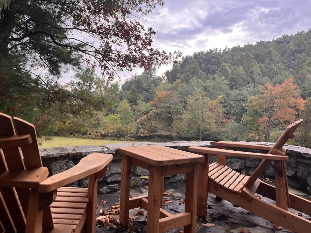 A wooden deck with Adirondack chairs and a table overlooking a forested hillside.