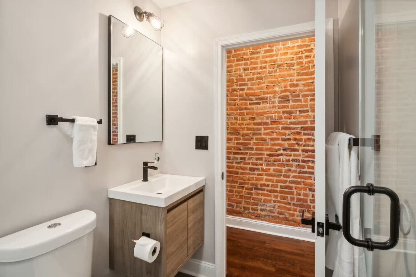 Een moderne badkamer met een wit wastafelmeubel, een spiegel, een toilet en een douchegedeelte met een baksteenmuur.