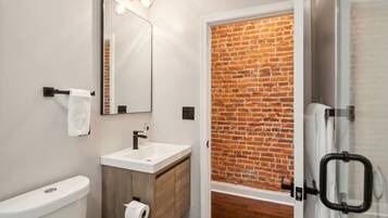 Een moderne badkamer met een wit wastafelmeubel, een spiegel, een toilet en een douchegedeelte met een baksteenmuur.