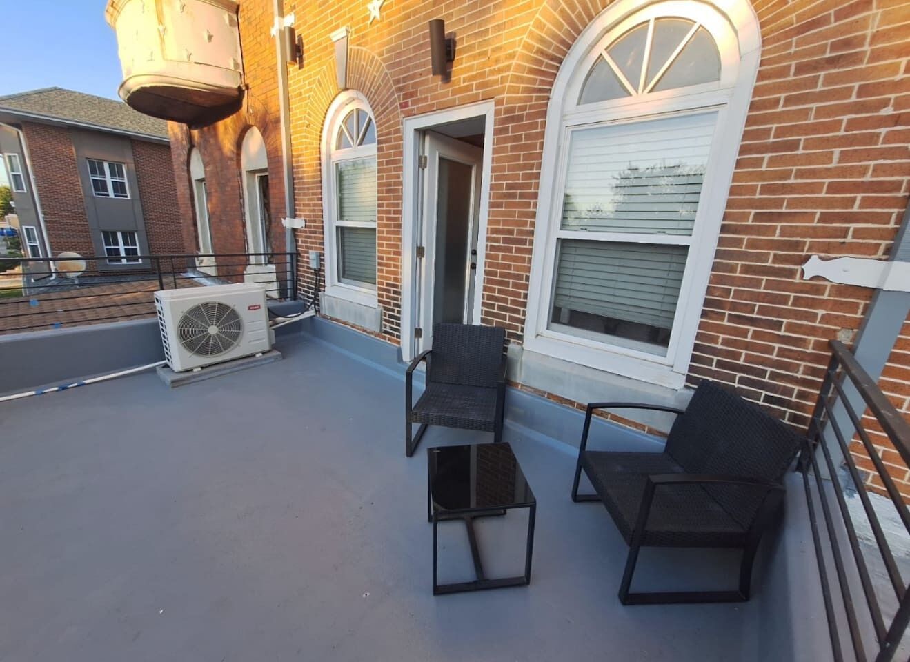 Eine Dachterrasse mit zwei Stühlen, einem kleinen Tisch, einem Wassertank und einem Backsteinhaus mit Rundbogenfenstern.