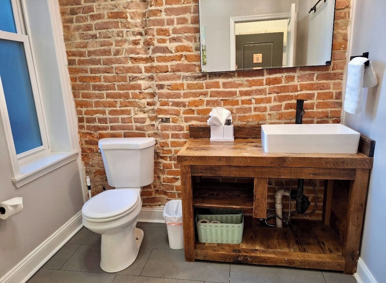 A bathroom with a brick wall, a white toilet, a wooden vanity with a sink, and a mirror above the vanity.