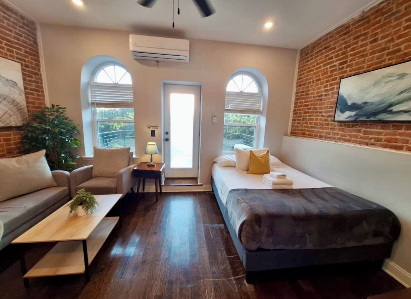 A modern room with a sofa, a bed, a coffee table, and a desk. There are two windows with blinds, a door, and an air conditioning unit on the wall.