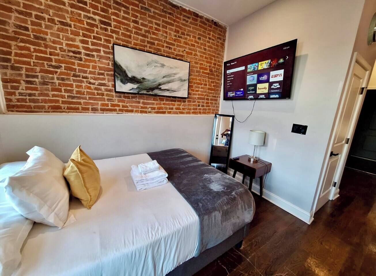 Ein Hotelzimmer mit Backsteinwand, einem flachbildfernseher über dem Bett, einem Schreibtisch mit Lampe und einer Tür zu einem weiteren Raum.