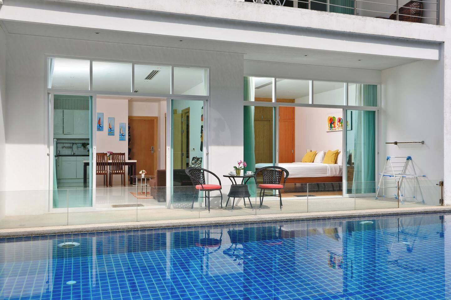 A modern hotel room with a glass wall, a bed, a small table with chairs, and a pool.