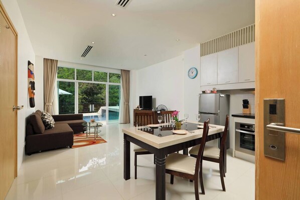 A modern living room with a sofa, dining table, and chairs, a kitchen with appliances, and a large window with a view of greenery.