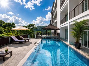 A swimming pool with lounge chairs and a gazebo, surrounded by a building with large windows and a view of greenery. - Suite Cascade 1br | Family deluxe | kamala falls (Tambon Kammala)