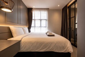 A modern hotel room with a large bed, a bedside table, a lamp, and a view of the outdoors through the window.