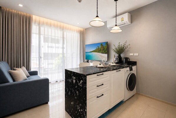 A modern living room with a sofa, a kitchen island with a washing machine, and a large window with curtains.