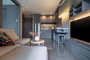 Una sala moderna con un sofá, una mesa de centro con objetos decorativos, un televisor de pantalla plana y una zona de cocina con un refrigerador y taburetes de bar.
