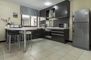 A modern kitchen with a stainless steel refrigerator, a microwave, and a built-in oven. There are two bar stools and a white countertop with a blender and a kettle.