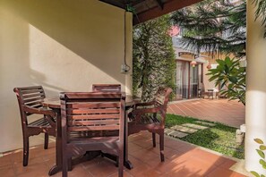 Een terras buiten met een houten tafel en stoelen, omgeven door een tuin en een gebouw met grote ramen.