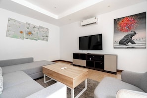 A modern living room with a flat-screen TV, grey sofas, a wooden coffee table, and artwork on the walls. - Villa Elara Bang Tao 3br | pool | east residence (Thep Krasatti)