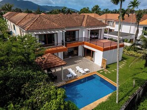 A two-story house with a swimming pool, surrounded by lush greenery and palm trees.