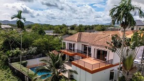 Una casa de dos pisos con piscina, rodeada de exuberante vegetación y palmeras.