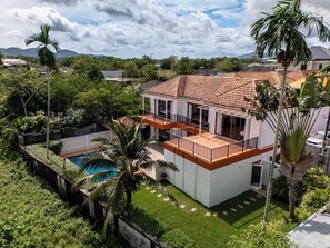 A two-story house with a swimming pool, surrounded by lush greenery and palm trees.