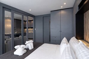 A modern bedroom with a large bed, a wardrobe, and a seating area with a view of the kitchen.