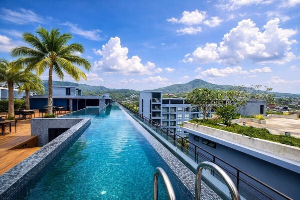 A swimming pool with a view of a mountain range, surrounded by modern buildings and palm trees. - Suite Zenith 1br|rooftop Pool &bar|citygate kamala (Tambon Kammala)