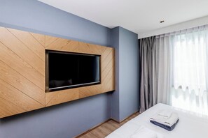 A hotel room with a large wall-mounted TV, a bed with neatly folded towels, and a window with sheer curtains. - Suite Elio 1br | Pools|gym & shuttle to nai harn (Rawai)