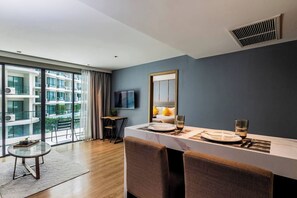 A modern hotel room with a dining area, a large window with a view of the city, a flat-screen TV mounted on the wall, and a ceiling vent.