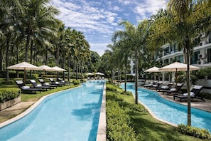 A swimming pool with lounge chairs and umbrellas, surrounded by palm trees and a building in the background. - Suite Elio 1br | Pools|gym & shuttle to nai harn (Rawai)