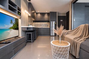 A modern kitchen with a large flat-screen TV, a refrigerator, and a dining area with stools. - Vista 1br | discounted stay | Citygate Kamala (Tambon Kammala)
