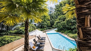Piscina com espreguiçadeiras e guarda-sóis, rodeada por vegetação exuberante e um edifício moderno ao fundo.