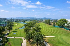 Een golfbaan met een waterhindernis, palmbomen en een verre weids uitzicht op een stad.