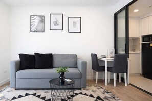 A modern living room with a grey sofa, black cushions, a black coffee table, and a patterned rug. There is a dining area with a white table and black chairs. Three framed pictures are hung on the wall above the sofa. - Suite Sky 1br | Rooftop pool & gym|laguna skypark (Thep Krasatti)