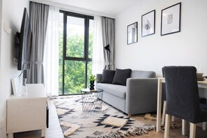 A modern living room with a grey sofa, a small round coffee table, a black chair, and a large window with curtains. - Suite Sky 1br | Rooftop pool & gym|laguna skypark (Thep Krasatti)