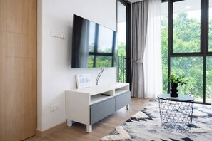 Una sala moderna con un televisor de pantalla plana montado en la pared, una mesita blanca con un vaso y vistas a la vegetación a través de grandes ventanales.