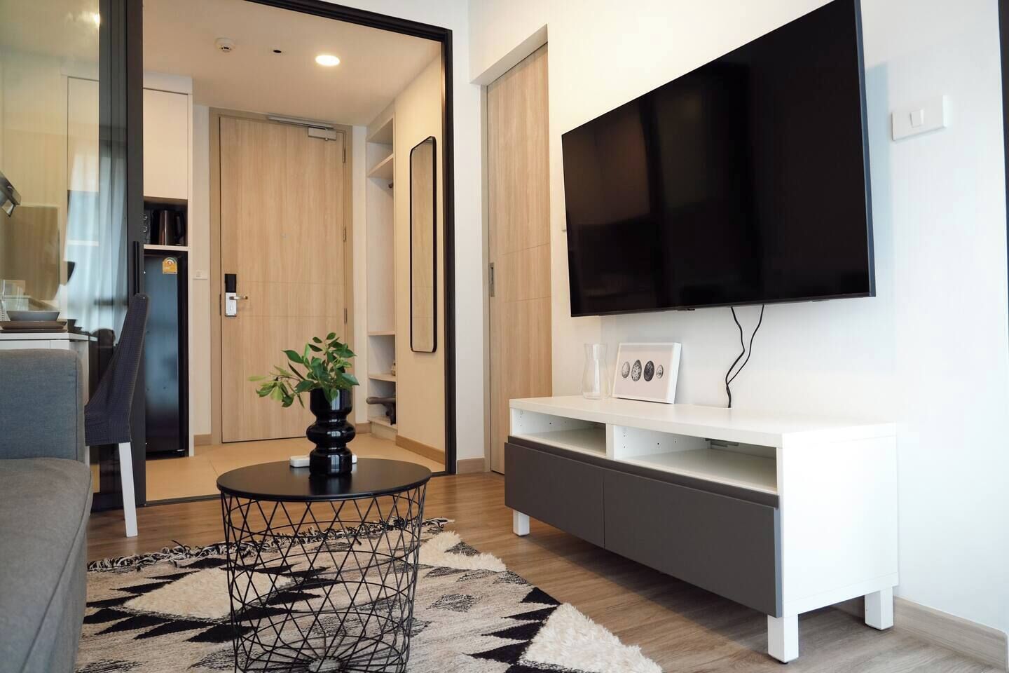 A modern living room with a flat-screen TV, a white entertainment unit, a round coffee table with a plant, and a patterned rug.