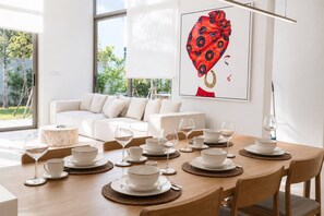 A modern dining area with a wooden table set for a meal, a white sofa in the background, and a large window providing a view of greenery outside.