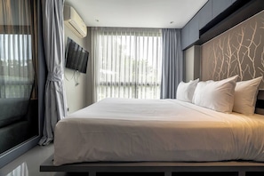 A modern hotel room with a large bed, a flat-screen TV mounted on the wall, and a view of the outdoors through sheer curtains.