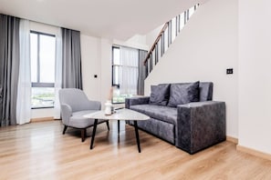 Ein modernes Wohnzimmer mit einem grauen Sofa, einem grauen Sessel und einem kleinen weißen Tisch. Rechts befindet sich eine Treppe und große Fenster mit Vorhängen.