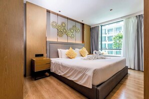 A hotel room with a large bed, a bedside table, a wall-mounted light fixture, and a view of the outside. - Suite Aluna 1br | Pools & shuttle to nai harn (Rawai)