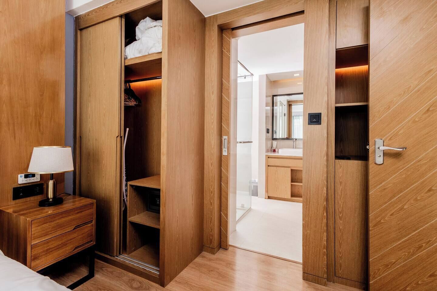 A hotel room with a wooden wardrobe, a bedside table with a lamp, and a view into the bathroom.