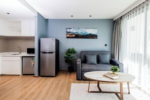 A modern apartment with a kitchen, a grey sofa, a round coffee table, and a large window with curtains. - Suite Nami 1br | Pools|gyms & shuttle to nai harn (Rawai)
