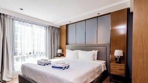A modern hotel room with a large bed, a wooden headboard, two bedside tables with lamps, and a view of the city through the window.