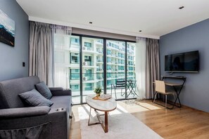 A modern living room with a grey sofa, a small round table, a TV, and a balcony with a view of a building.