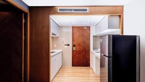 Un pasillo estrecho con una puerta de madera a la izquierda, un refrigerador negro a la derecha y una encimera blanca con un lavamanos en el centro.