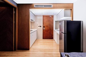 Un pasillo estrecho con una puerta de madera a la izquierda, un refrigerador negro a la derecha y una encimera blanca con un lavamanos en el centro.