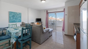 Espaço de estar com sofá, mesa de jantar, cadeiras, televisão e varanda com vista.