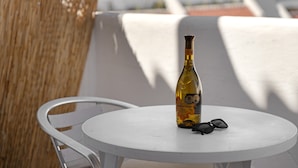 Uma garrafa de vinho sobre uma mesa branca, com um par de óculos de sol ao lado, colocada numa varanda com uma cadeira e outra cadeira branca ao fundo.