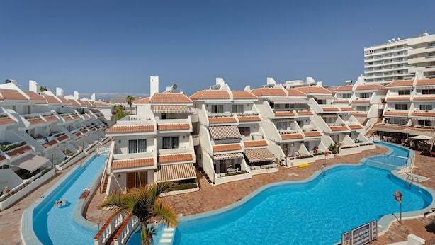Resort com vários edifícios brancos, uma piscina central e céu azul límpido.