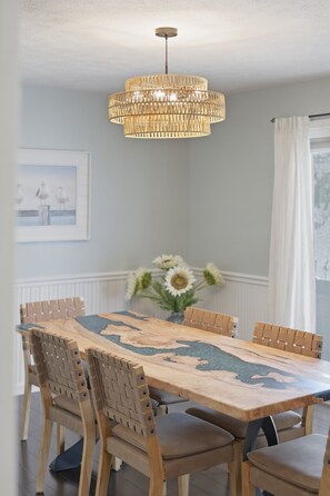 Uma sala de jantar com mesa de madeira, cadeiras, luminária pendente e quadro na parede.