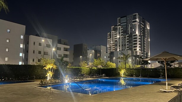 A nighttime swimming pool area with a fountain, surrounded by buildings and a well-lit patio area.