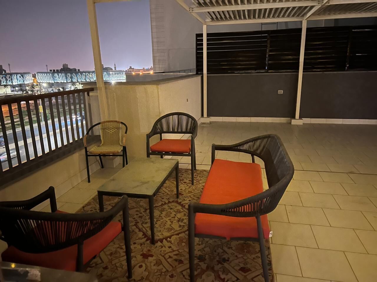 A balcony with a table and chairs, a patterned rug, and a view of the cityscape at dusk.