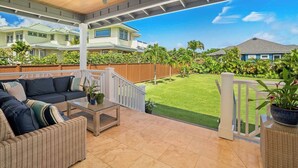 A covered patio with wicker furniture, a glass coffee table, and a view of a well-maintained lawn and neighboring houses.